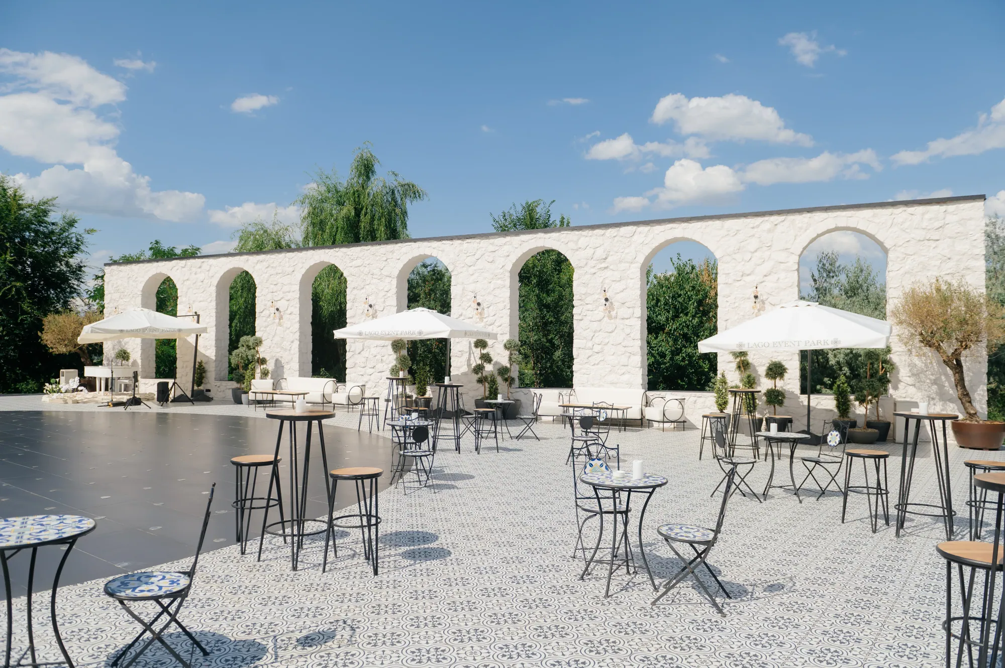 Curte cu arcade din piatră albă, umbrele și mese înalte de cocktail