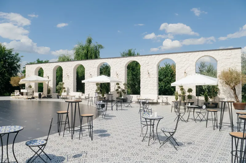 Curte cu arcade din piatră albă, umbrele și mese înalte de cocktail